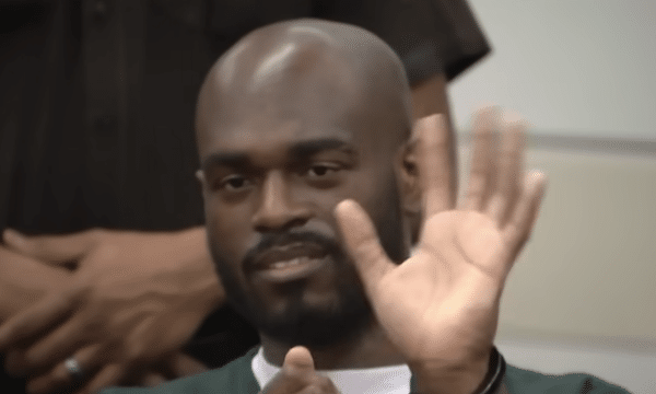 Close-up of a bald man with a beard seated in a courtroom, wearing dark green clothing and looking slightly to the side.