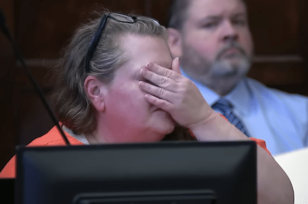 Woman in an orange jail uniform covers her mouth with one hand while seated in a courtroom, with a blurred man in the background.