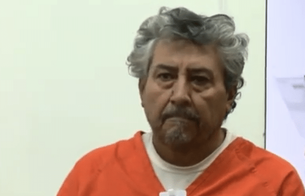 A gray-haired man in an orange jail uniform looks forward during what appears to be a courtroom hearing.