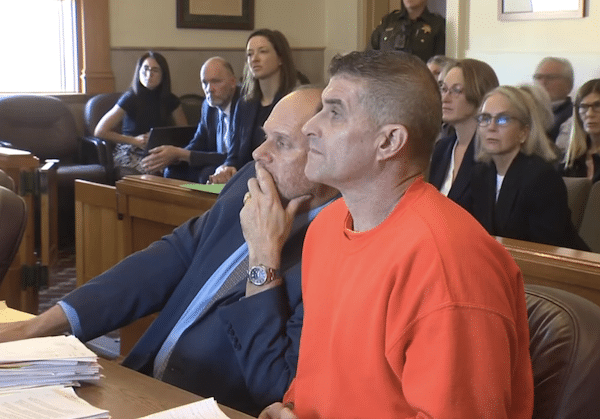 Courtroom photo of former MLB pitcher Daniel Serafini in a bright orange jail shirt seated beside his attorney at the defense table, with deputies, spectators and framed wall art visible behind them.