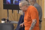 Older man in an orange jail uniform, with gray hair tied back and tattoos on his forearm, walks with a cane in a courtroom while another older man stands behind him and video screens display remote participants.