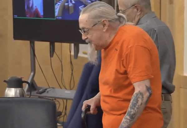 Older man in an orange jail uniform, with gray hair tied back and tattoos on his forearm, walks with a cane in a courtroom while another older man stands behind him and video screens display remote participants.