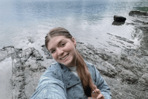 Young woman smiling for a selfie on rocky shoreline beside calm lake water.