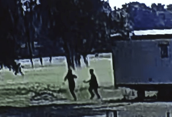 Surveillance footage showing two silhouetted figures in a grassy outdoor area, with trees and a large rectangular object nearby.