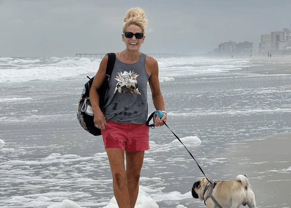 Woman in sunglasses walks along a windy beach holding a dog leash, wearing a gray tank top and bright pink shorts under a cloudy sky.