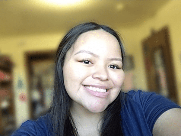 Smiling young woman with long dark hair taking a selfie indoors against a softly blurred background.