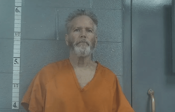 Mugshot of an older man with gray hair and a full gray beard wearing an orange jail shirt while standing against a height chart and gray cinder block wall.