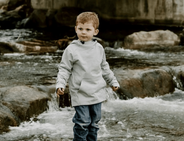Young child in a light gray hoodie and jeans stands in a shallow rocky stream, with rushing water and a blurred stone backdrop behind him.
