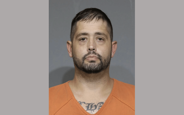 Front-facing booking photo of an adult man with short dark hair and a trimmed beard wearing an orange jail uniform against a gray background.