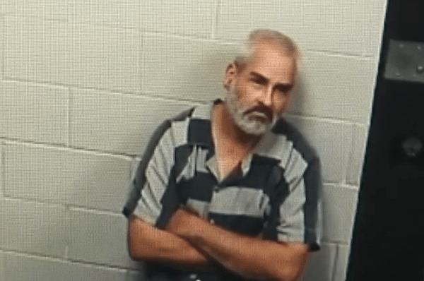Older man with short gray hair and a beard sits with his arms crossed in a sparse interview room with cinderblock walls.