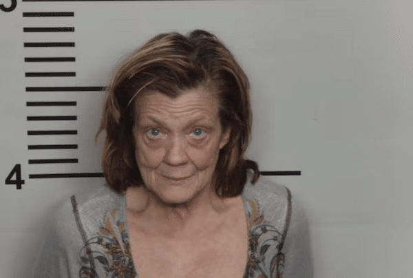 Mugshot-style image of a woman with shoulder-length brown hair standing in front of a height chart, wearing a light gray top and looking straight at the camera.