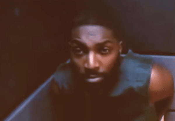 A bearded man with short hair sits in what appears to be a courtroom or holding area, leaning forward and looking toward the camera in a low-resolution image.