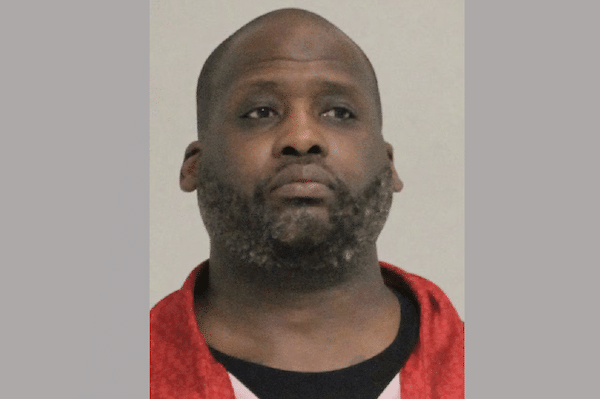 Portrait of a serious-looking man with a shaved head and short beard wearing a red garment against a plain light background.