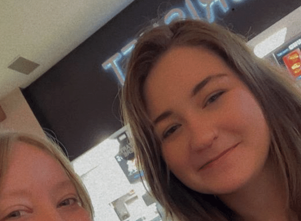 Selfie-style photo shows two young women smiling inside a business with a bright sign behind them.