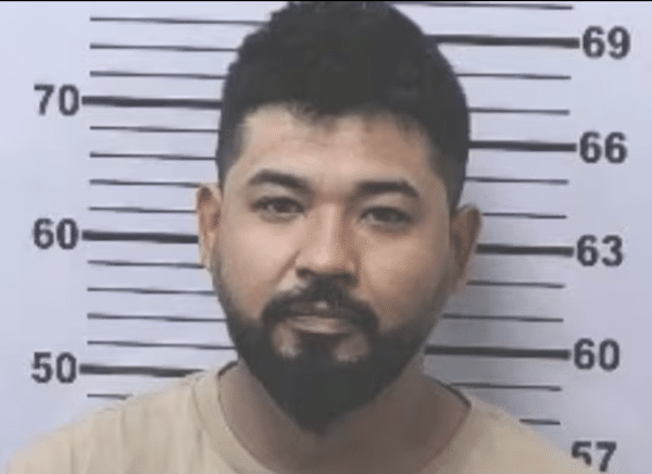 Mugshot-style booking photo of a dark-haired man with a beard and mustache wearing a light tan shirt in front of a height chart.