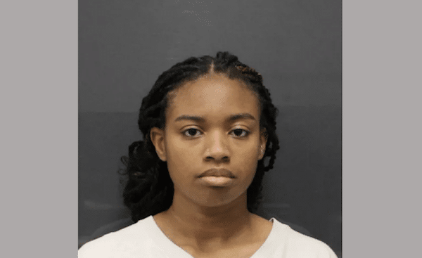 Front-facing portrait of a young woman with braided hair, neutral expression, and a light gray shirt against a dark background.