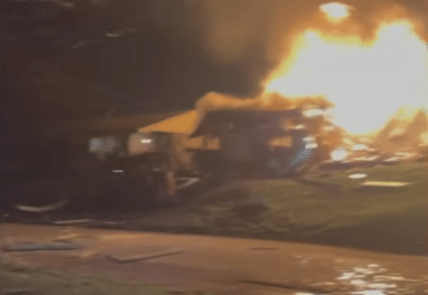A house burns intensely at night as flames shoot high above the roof and debris glows across the yard.