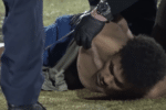 A person lies on the grass while gloved hands from officers hold their upper body during an outdoor arrest.