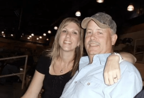 A smiling woman with her arm around an older man in a baseball cap as they pose together indoors under dim hanging lights.