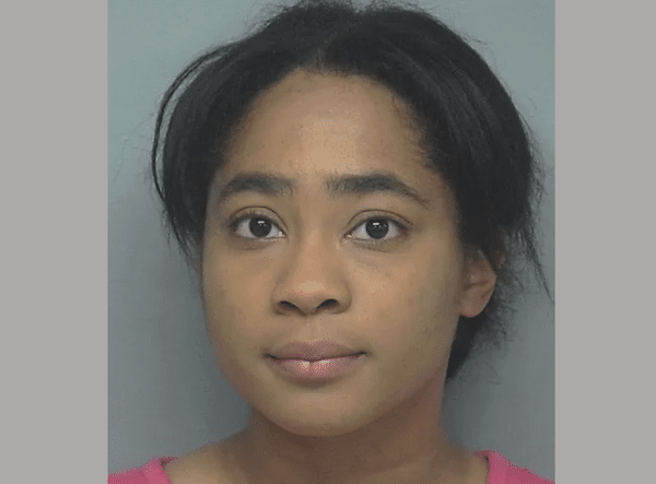 Mugshot-style portrait of a woman facing forward against a gray background, wearing a pink top with dark hair loosely pulled back and a neutral expression.