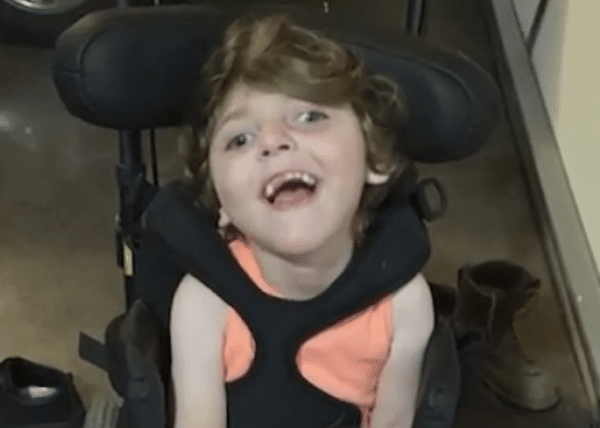 Smiling child with short brown hair sits in a supportive wheelchair wearing a bright coral sleeveless shirt indoors.