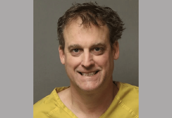 Booking-style photo shows a man in a yellow jail uniform facing the camera against a gray background.