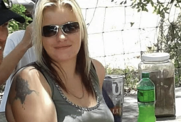 Woman with blonde and dark hair wearing sunglasses and a sleeveless top, seated outdoors at a table with a green soda bottle and jars nearby.