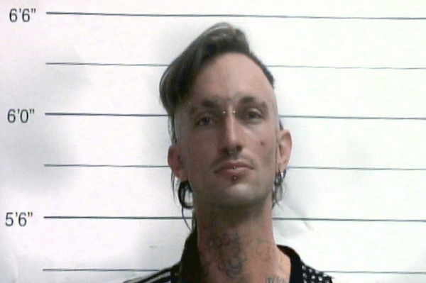 Front-facing portrait of a man with shaved sides, longer dark hair on top, and facial piercings against a height chart background.