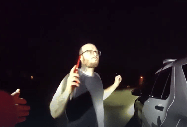 A man wearing glasses stands outside at night with his hands raised as another person reaches toward him in bright flashlight glare.