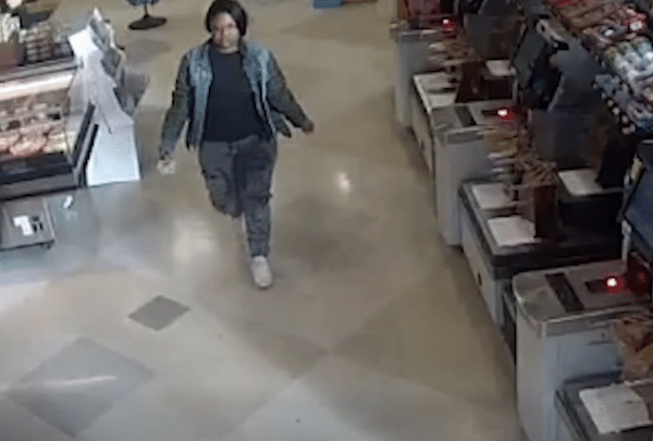 Surveillance-style image of a person walking through a retail store aisle, wearing a dark shirt, green jacket, jeans and white sneakers, with checkout stations lining one side.