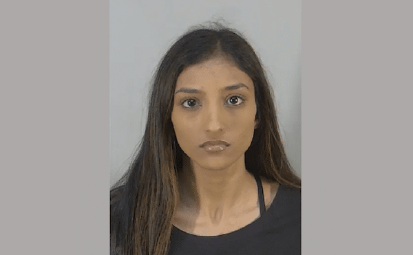 Mugshot-style portrait of a woman with long dark hair, wearing a black top, facing the camera against a light gray background.