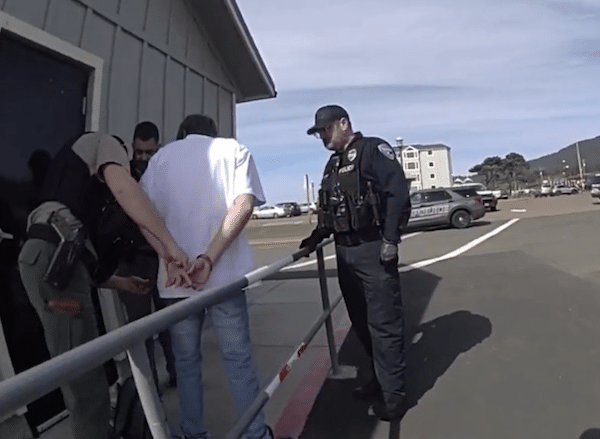 Police officers detain a person with hands behind their back outside a building near a parking area.