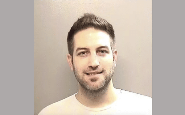 Head-and-shoulders portrait of a man with short dark hair and light stubble smiling slightly against a plain beige wall.