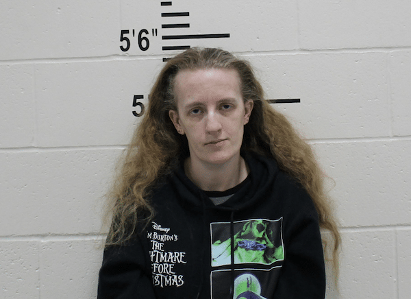 Mugshot-style photo of a woman with long light brown hair wearing a black hoodie, standing in front of a height chart on a white cinderblock wall.