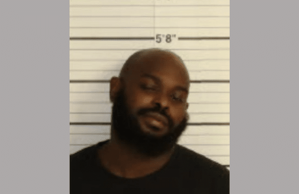 Mugshot-style photo of a bald man with a beard in a dark shirt standing in front of a height chart.