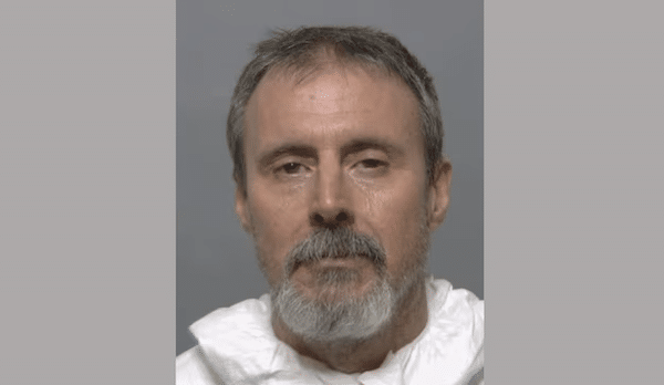 Booking-style photo of an older man with short graying hair, a gray beard and a white garment against a plain gray background.