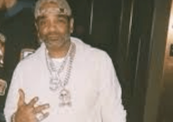 Blurry photo of a man wearing a light hoodie, chain necklaces and a patterned baseball cap, posing indoors with one hand raised.