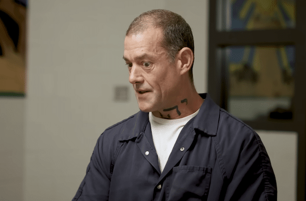 Man in a navy jail uniform with a neck tattoo looks to the side during an indoor interview or court related setting.