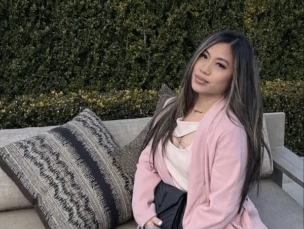 A woman with long dark hair sits on an outdoor couch, wearing a pink cardigan over a light top and dark skirt, with patterned pillows beside her and a hedge in the background.