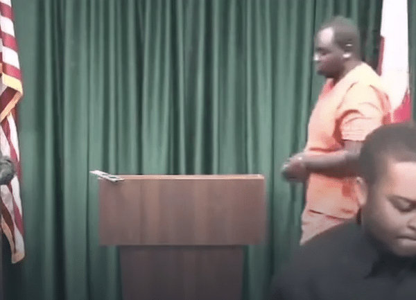 A man in orange jail clothing stands at a courtroom podium beside a uniformed officer and an American flag.