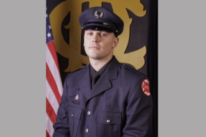 Portrait of a young firefighter in a dark navy dress uniform and cap standing beside a U.S. flag.