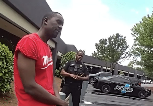 A man in a red T-shirt stands near a police officer in a parking lot, with a marked police car and office buildings in the background.