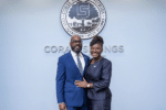Two people in formal business attire stand smiling beneath the City of Coral Springs, Florida seal on a blue wall.