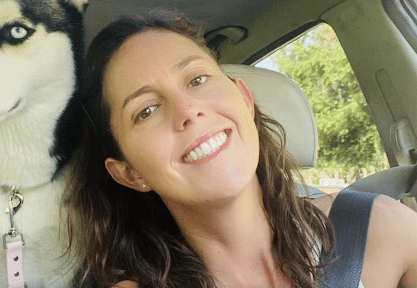 Smiling woman taking a car selfie with a black-and-white husky sitting beside her.