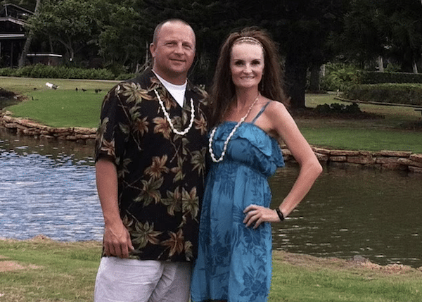 Man and woman wearing leis pose together on grass beside a pond in a park-like setting.