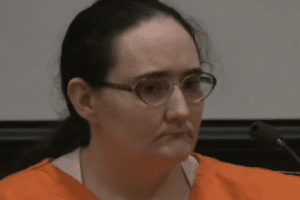 Person in an orange jail uniform sits at a courtroom microphone during a hearing.