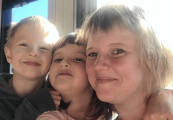 A woman smiles for a close-up selfie with two young children beside a bright window.