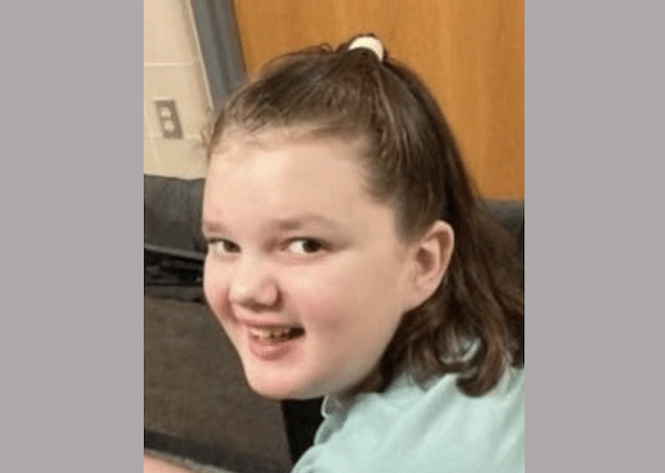 Smiling child with a ponytail looks toward the camera while seated indoors.