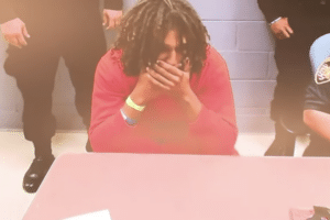 A man in a red sweatshirt sits at a table with his hands near his face while officers stand nearby.