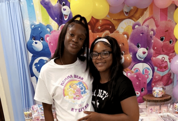 Two people pose together in front of a colorful cartoon bear backdrop and party decorations.